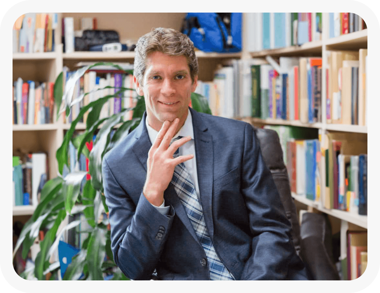 Dr. Lundell looking at the camera and smiling. In his office surrounded by books, in a suit.